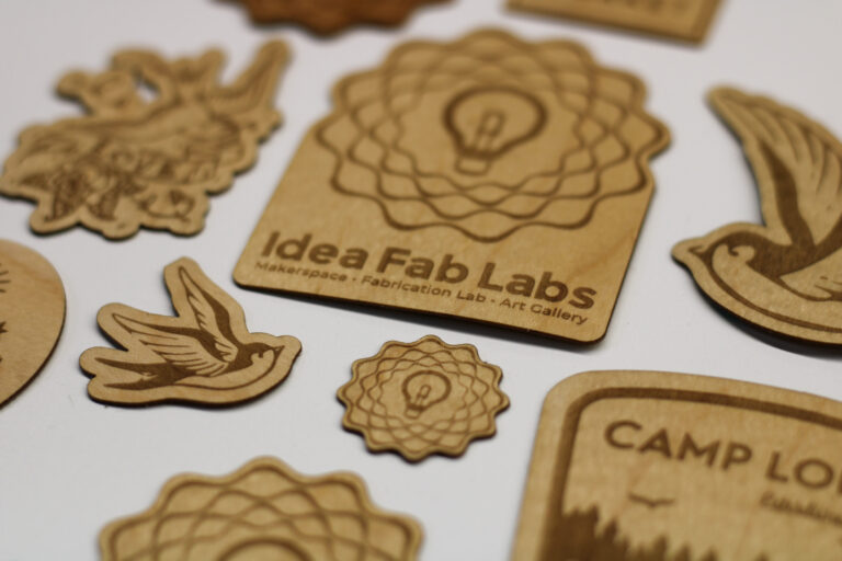 An array of wooden stickers with various carvings such as a lightbulb design and birds in flight, with 'Idea Fab Labs' and other texts etched onto them, laid out on a white background.